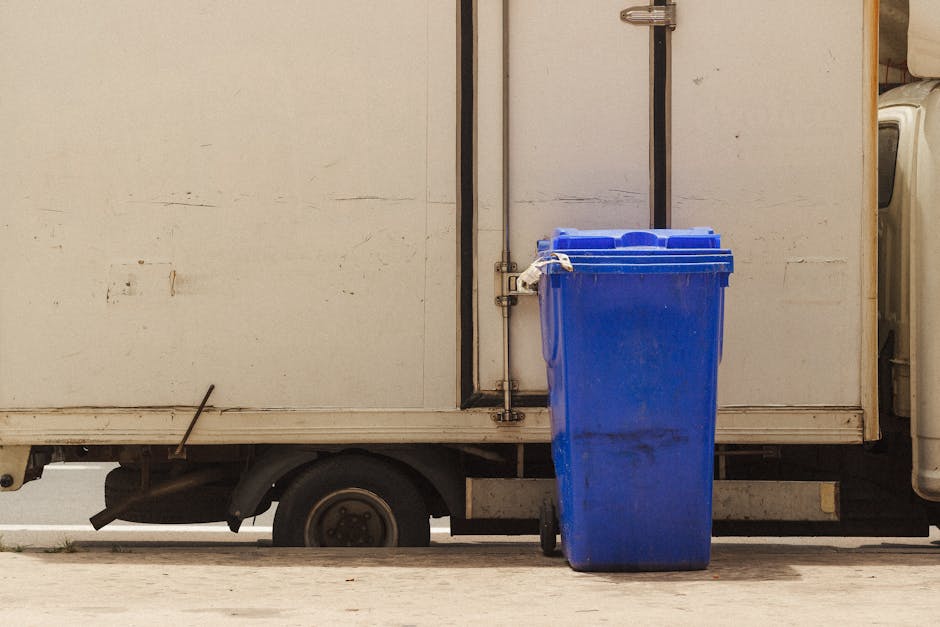A bright blue wheeled trash bin with a closed lid is positioned next to the side of a small commercial vehicle or van, which has a plain white, slightly weathered exterior. The bin is made of sturdy p