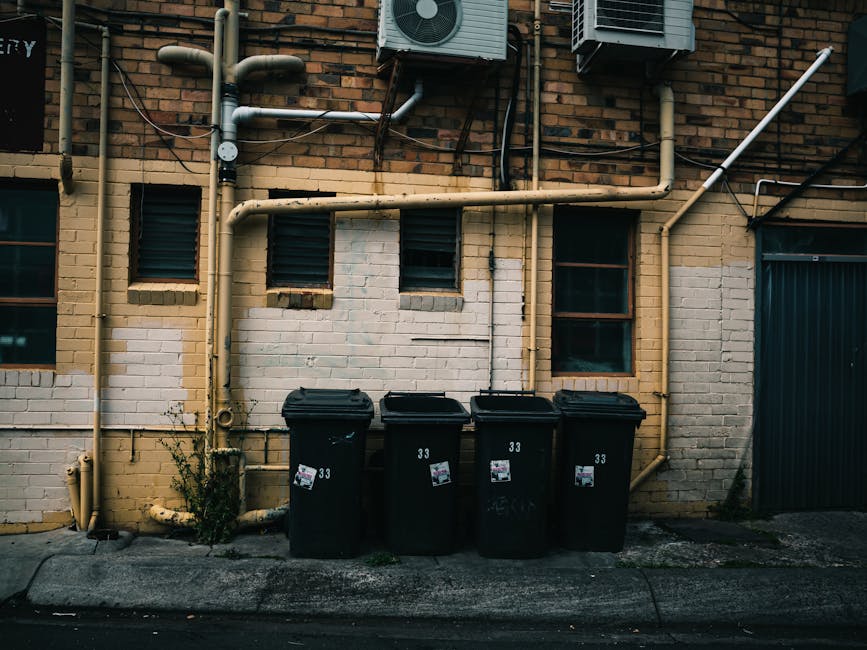 A bright blue wheeled trash bin with a closed lid is positioned next to the side of a small commercial vehicle or van, which has a plain white, slightly weathered exterior. The bin is made of sturdy p